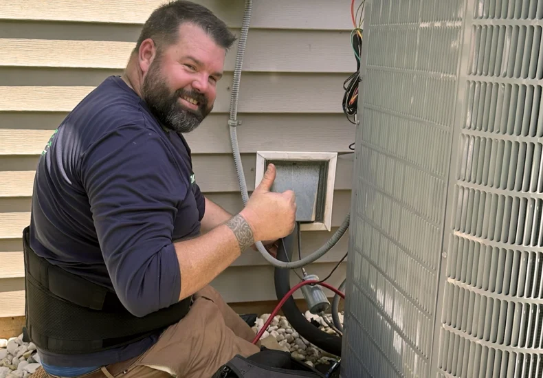Technician kneeling and giving thumbs up while working on an outdoor HVAC unit next to house siding and vent.