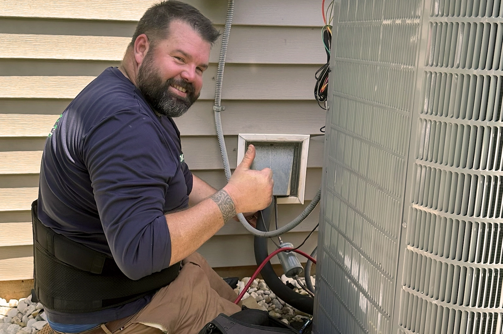 Technician kneeling and giving thumbs up while working on an outdoor HVAC unit next to house siding and vent.